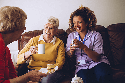 Elderly women talking over tea with a younger female carer sitting with them on a sofa in a care home