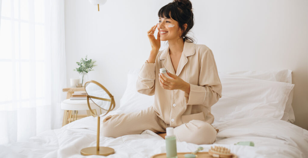 Happy woman doing routine skin care at home with beauty products. Woman sitting on bed at home and applying face cream.