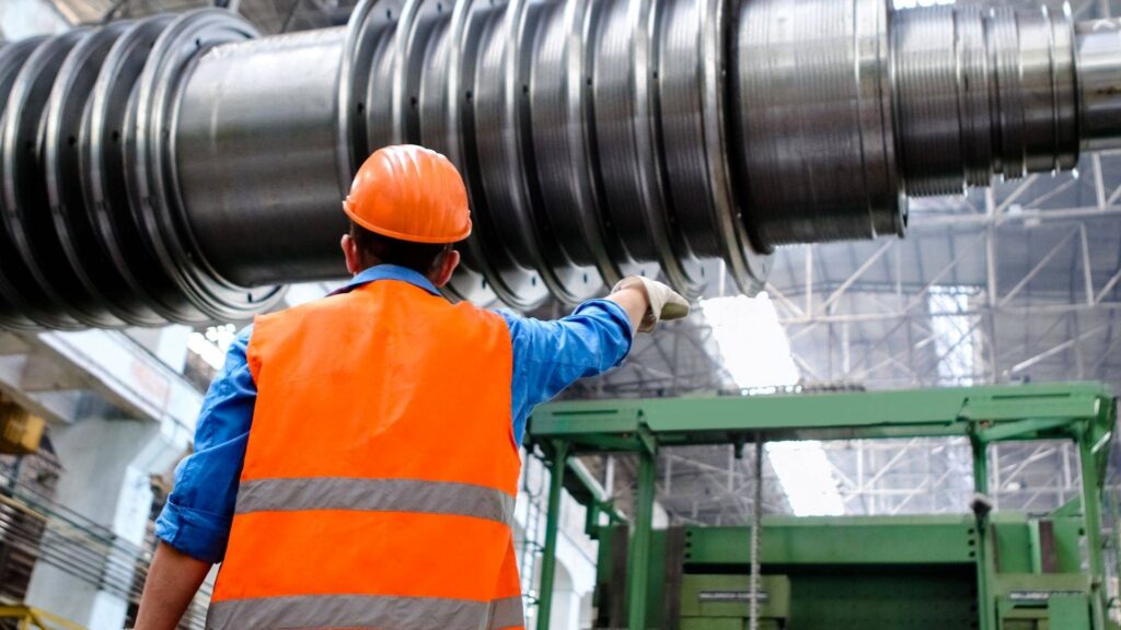 A worker in a safety vest and hard hat points toward a large metal industrial machine component in a factory