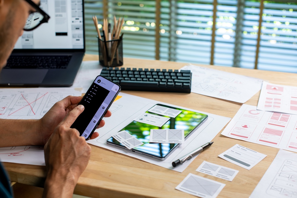 Mobile App Development - UX UI designer testing prototype on a phone, working on wireframes for a website and mobile application at a desk