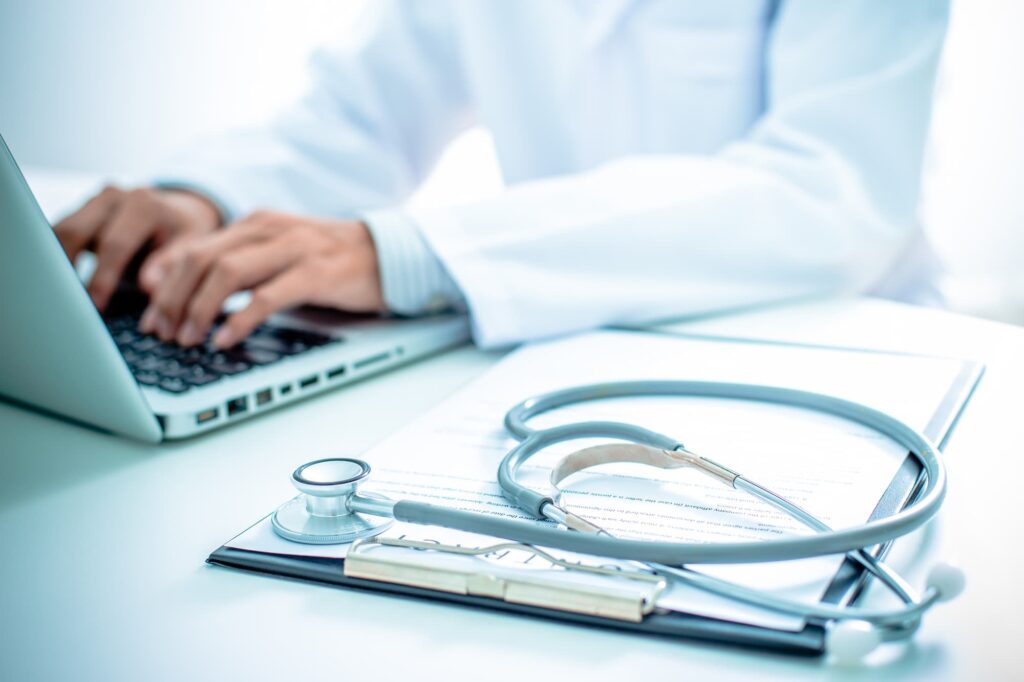 Health insights - Alt text: Doctor in white coat typing on a laptop with a stethoscope and clipboard on the desk.