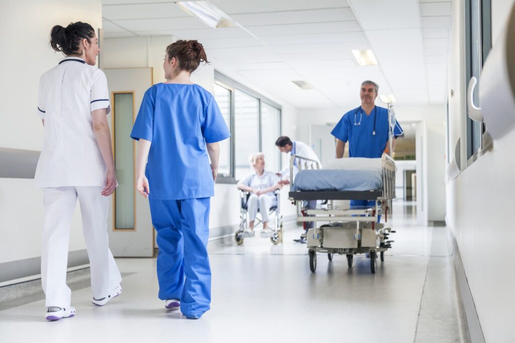Healthcare data analytics - Young female nurses walk down a hospital hallway while a doctor pushes a hospital bed and another nurse assists an old women in a wheelchair.

