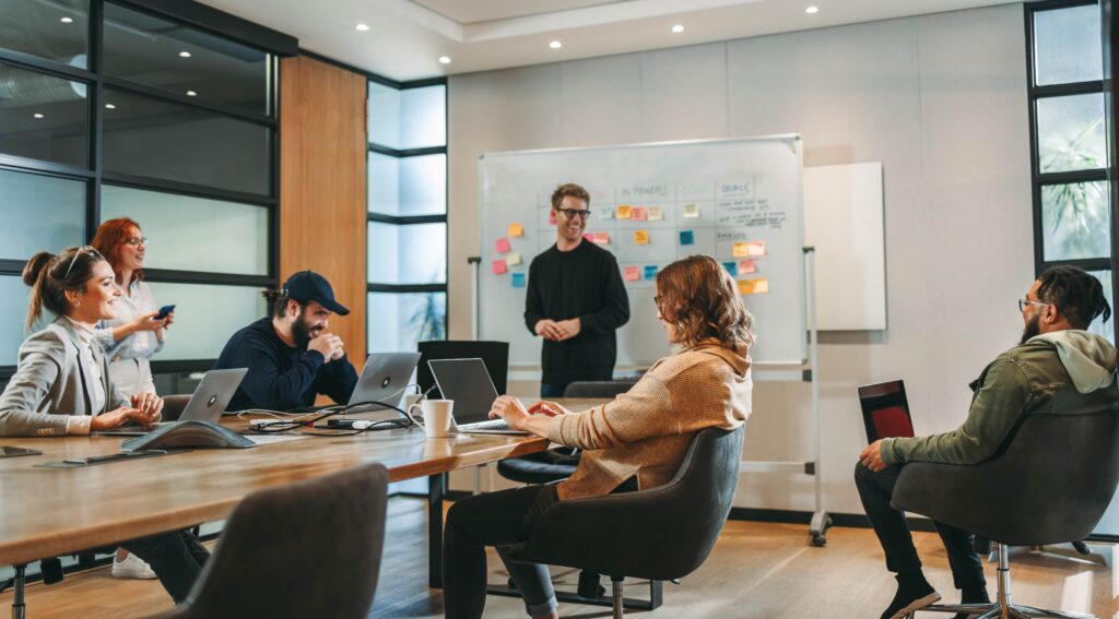 UX and digital product design - A group of people in a modern office, with one person presenting at a whiteboard and others seated with laptops.

