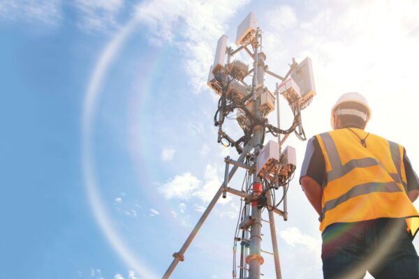 Helmeted male engineer works in the field with a telecommunication tower that controls cellular electrical installations to inspect and maintain 5G networks installed on high-rise buildings