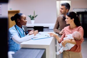 Oxford Health NHS Foundation Trust - Couple with a baby talking to a hospital receptionist sitting behind a reception desk