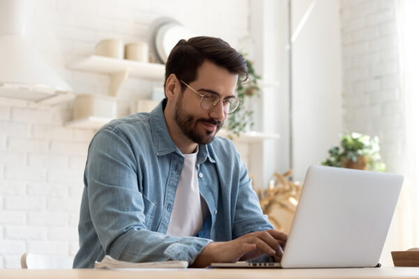 Focused young man wearing glasses using laptop, typing on keyboard