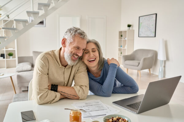 Smiling middle aged senior 50s husband and wife having fun satisfied with buying insurance, paying bills on their laptop.