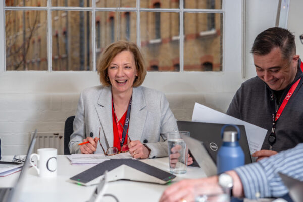 People in a meeting room sitting at a table with laptops, documents, and drinks in front of large windows.