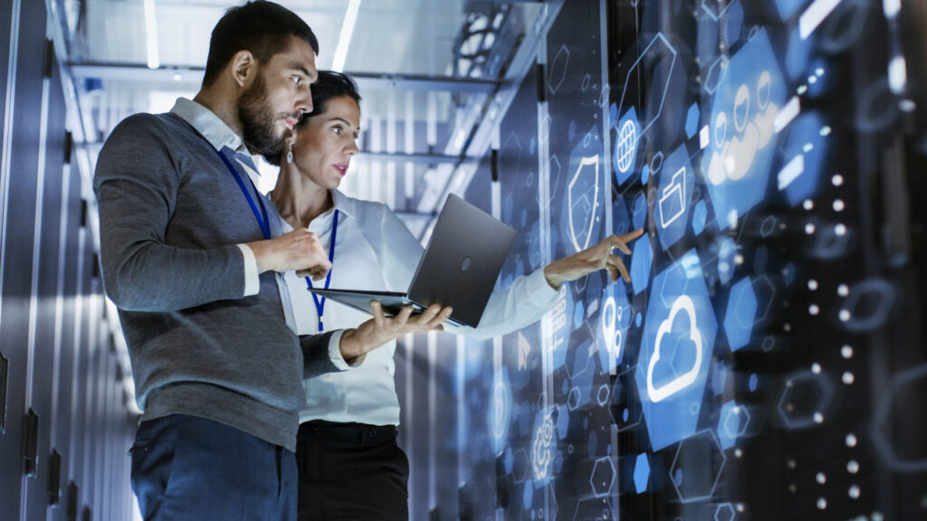 Male IT Specialist Holds Laptop and Discusses Work with Female Server Technician, they are standing in a server room together